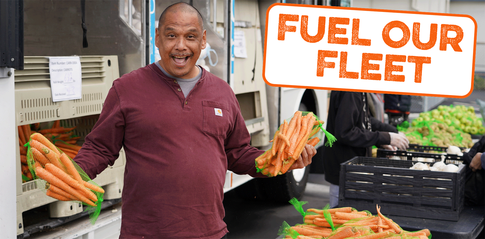 Man holding carrots in front of a Food Bank vehicle with sign that reads Fuel Our Fleet